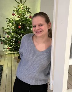 Cora Peterson lener seg til vegg og smiler, foran juletreet.