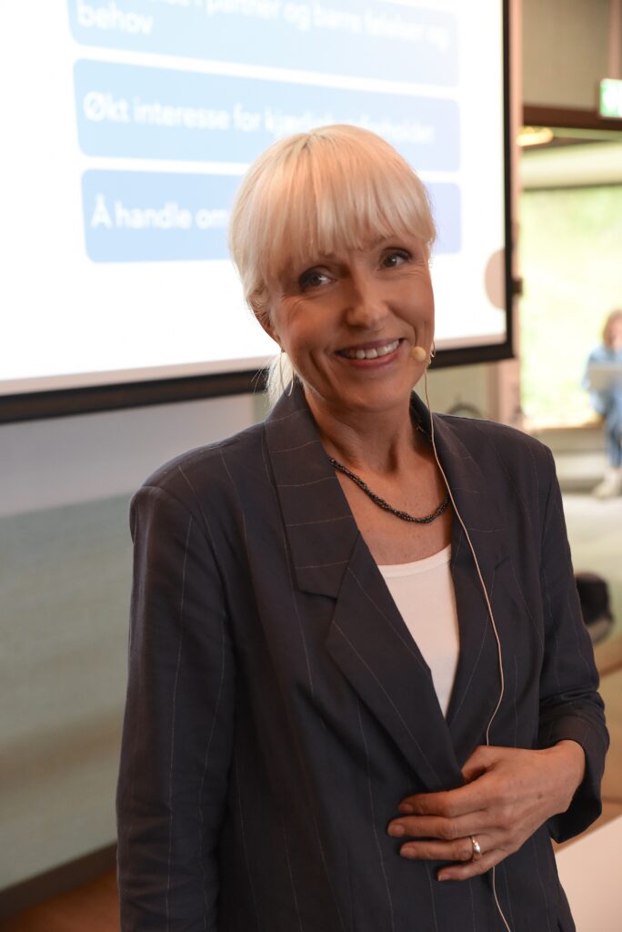 Ingunn Rangul Askeland smiler til kamera foran skjerm med foredrag.
